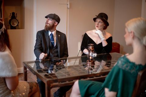 Eine Gruppe von elegant gekleideten Personen sitzt an einem Tisch in einem stilvollen Raum. Die Männer tragen Anzüge und Hüte, während die Frauen sich in klassischer Abendgarderobe präsentieren.