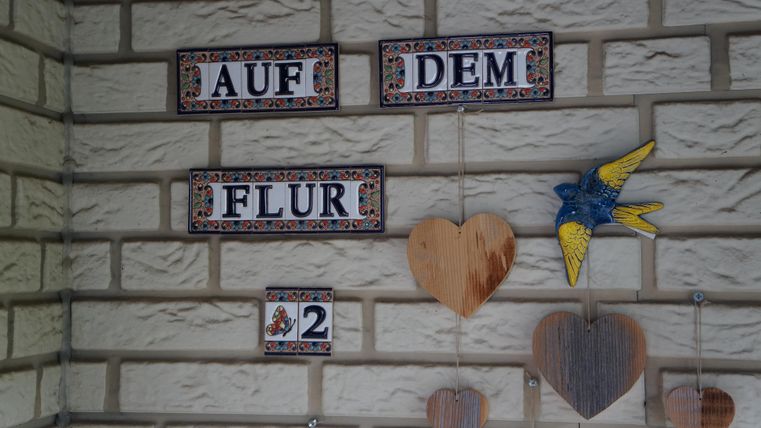 Eine Wand mit dekorativen Schildern, die "Auf dem Flur 2" zeigen. Daneben hängen Holzherzen und eine bunte Vogelfigur.