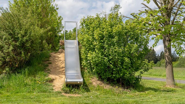 Eine Rutsche aus Metall steht in einem grünen Park. Umgeben von Büschen und Bäumen, lädt sie zum Spielen ein.