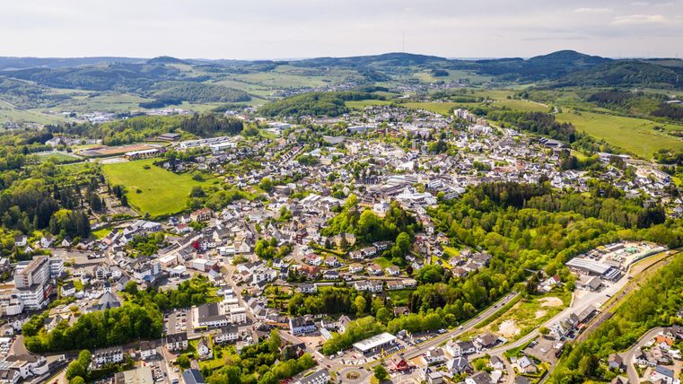 Eine luftige Ansicht einer kleinen Stadt umgeben von grünen Hügeln und Wäldern. Die Stadt hat zahlreiche Gebäude und Straßen, die sich harmonisch in die Landschaft einfügen.