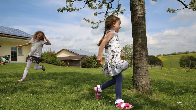 Zwei Kinder spielen fröhlich im Gras unter einem Baum. Im Hintergrund sind ein Haus und eine grüne Landschaft zu sehen.