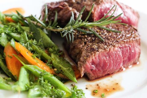 Ein saftiges Stück Rindersteak mit Gewürzen und frischem Rosmarin, serviert auf einem Teller. Daneben liegen bunte Gemüsesorten wie grüne Bohnen und Karotten.