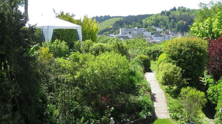 Ein schöner Garten mit üppigem Grün und bunten Pflanzen. Im Hintergrund ist eine malerische Landschaft mit Häusern und hügeligen Wäldern zu sehen.