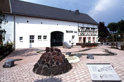 Ein historisches Gebäude mit einem modernen Eingang und einer malerischen Umgebung. Vor dem Gebäude befindet sich eine künstlerische Landschaftsgestaltung mit Steinen und Vulkanformationen.