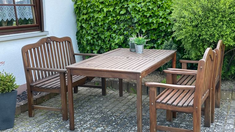 Ein Holztablett mit vier Stühlen steht im Freien. Im Hintergrund sind grüne Sträucher zu sehen.