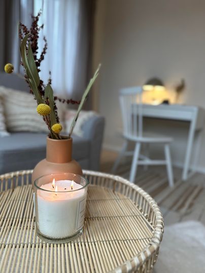 Eine gemütliche Wohnatmosphäre mit einer Kerze und einer Vase auf einem Tisch. Im Hintergrund ist ein helles Zimmer mit einem Schreibtisch und einem Stuhl sichtbar.