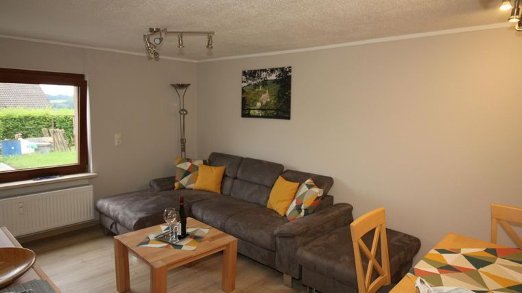 Ein gemütliches Wohnzimmer mit einer grauen Couch und bunten Kissen. Der Raum ist hell und einladend, mit einem Tisch und einer Pflanze.