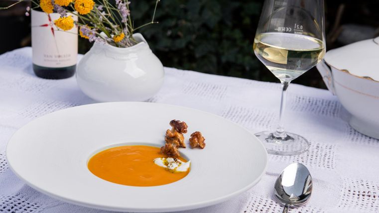 Eine elegante Suppe in einem weißen Teller, garniert mit Eifelfrische und einem Klecks Sahne. Daneben steht ein Glas Weißwein und eine kleine Blumenvase.