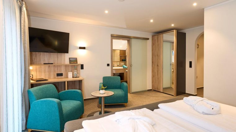 Ein modernes Hotelzimmer mit zwei Einzelbetten. Es gibt einen kleinen Wohnbereich mit blauen Sesseln und einem Tisch sowie einen Fernseher an der Wand.