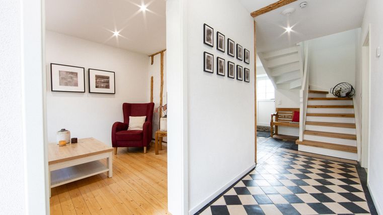 A bright hallway with a checkered floor and a staircase. On the left side, there is a red armchair and some pictures on the wall.