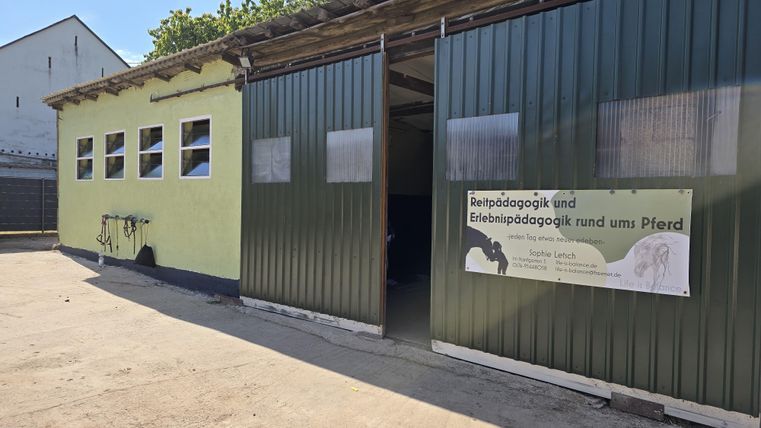 Ein ehemaliger Stall mit grünen Wänden und geöffneten Türen. An der Wand hängt ein Schild zur tiergestützten Pädagogik.