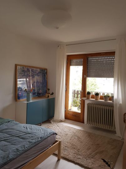 Ein helles Schlafzimmer mit einem Bett, einem Schreibtisch und großen Fenstern. Auf dem Fensterbrett stehen Pflanzen und der Raum wirkt freundlich und einladend.