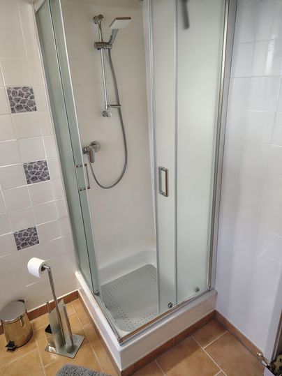 Eine moderne Dusche mit Glasfront und Handbrause. Der Boden ist gefliest und es gibt einen Handtuchhalter sowie eine Toilettenpapierrolle.