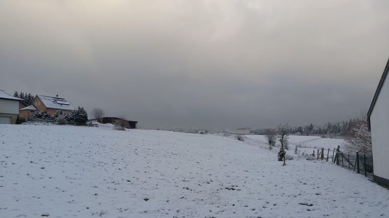 Eine verschneite Landschaft mit einem leicht bewölkten Himmel. Im Hintergrund sind einige Häuser und Bäume zu sehen.