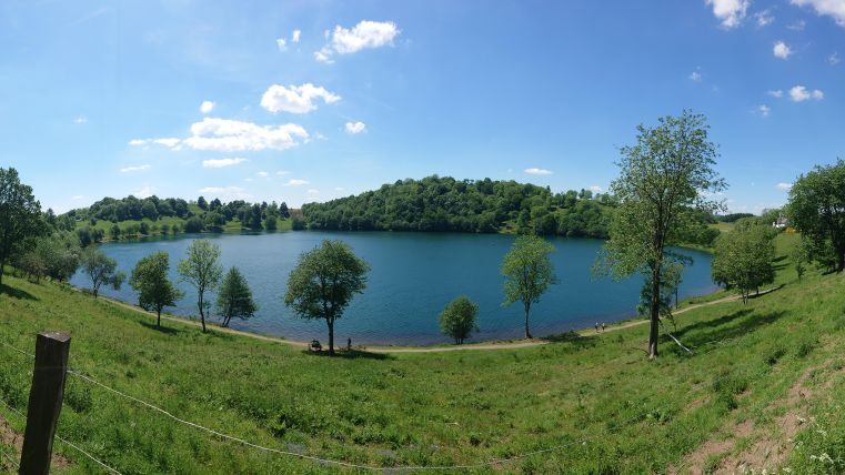 Ein ruhiger See umgeben von grünen Wiesen und Bäumen. Der Himmel ist klar und blau mit einigen weißen Wolken.