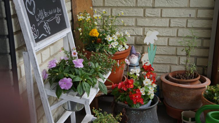 Ein schöner Balkon mit bunten Blumen in Töpfen. Im Hintergrund steht eine weiße Leiter mit einer Tafel.