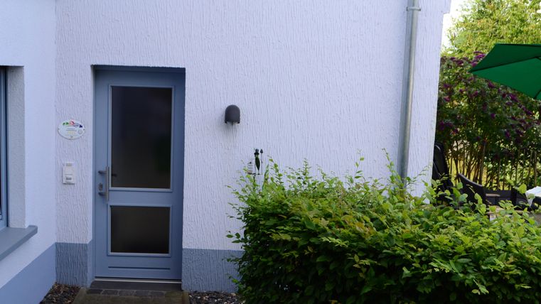 Ein Eingang zu einem Haus mit einer grauen Tür und einem kleinen Vorbereich. Neben der Tür ist eine grüne Hecke und ein Teil eines Gartens sichtbar.