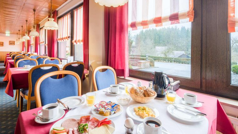 Ein einladender Frühstücksraum mit gedeckten Tischen und einer vielfältigen Auswahl an Speisen. Im Hintergrund sind Fenster mit rotem Vorhang und Blick auf die Landschaft zu sehen.