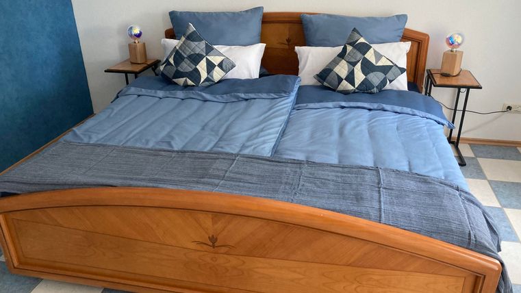 Ein gemütliches Schlafzimmer mit einem Doppelbett in Holzoptik. Die Bettwäsche ist blau und die Kissen haben diverse Muster.