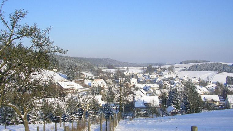 Eine verschneite Landschaft mit einem kleinen Dorf im Tal. Die Bäume und Hügel sind mit Schnee bedeckt und der Himmel ist klar und blau.
