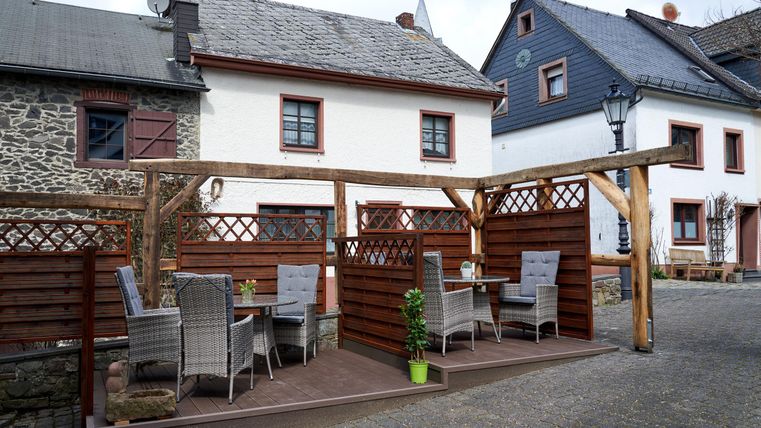 Ein gemütlicher Außenbereich mit Tischen und Stühlen aus Rattan. Im Hintergrund sind traditionelle Häuser zu sehen.