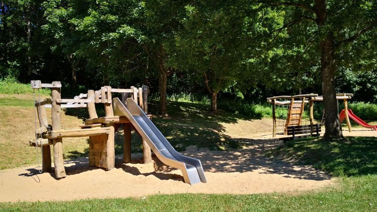 Ein Spielplatz mit einer Rutsche und einer Schaukel. Der Bereich ist von Bäumen umgeben und hat eine sandige Unterlage.