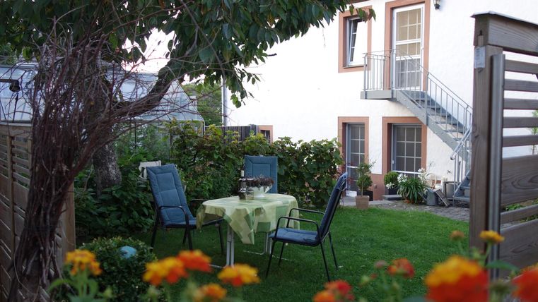 Ein gemütlicher Garten mit einem Tisch und Stühlen. Im Hintergrund ist ein Haus mit einem Balkon zu sehen.