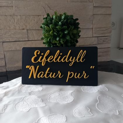 A sign with the inscription "Eifelidyll - Nature pure" stands on a table. In the background, a small plant can be seen.