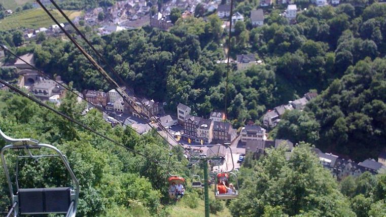 Eine Seilbahn fährt über eine grüne Hügel landschaft mit Blick auf ein kleines Dorf. Die Umgebung ist von Bäumen umgeben und zeigt eine malerische Aussicht.