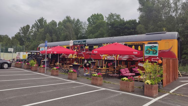 Ein bunt dekorierter Restaurantwagen mit roten Sonnenschirmen steht in einem Parkplatz. Umgeben von Pflanzen und Sitzgelegenheiten lädt er zum Verweilen ein.