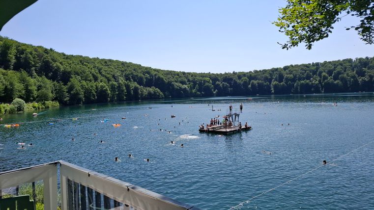 Ein malerischer See umgeben von grünen Wäldern. Menschen schwimmen und spielen auf einer Plattform im Wasser.