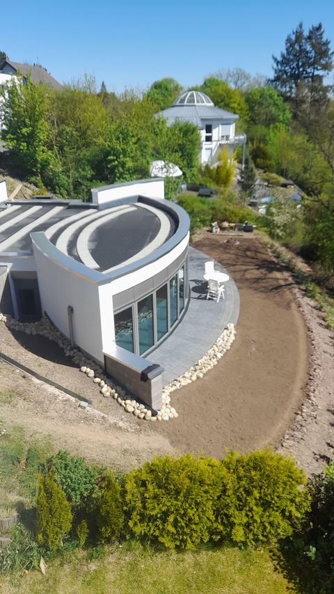 Ein modernes Haus mit einer geschwungenen Fassade und großen Fenstern. Umgeben von einer grünen Landschaft und einem angelegten Garten.