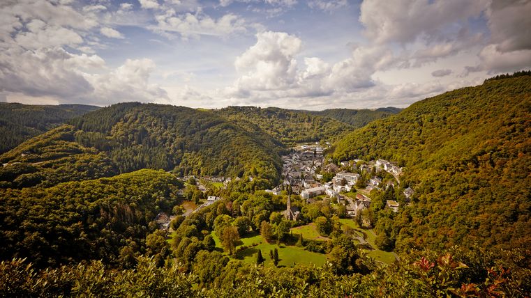 Panoramablick bei bewölktem Wetter auf den Ort Bad Bertrich, umgeben von einer hügeligen, dicht bewaldeten Landschaft.
