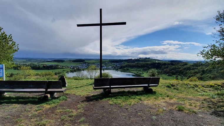 Aussichtspunkt mit einem großen Kreuz und Bänken. Im Hintergrund fließt ein Fluss unter einem klaren blauen Himmel.
