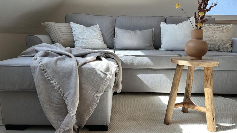 Ein gemütliches Wohnzimmer mit einer grauen Couch und mehreren Kissen. Neben der Couch steht ein Holztisch mit einer Vase und frischen Blumen.