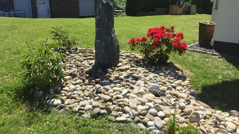 Ein gepflegter Garten mit einem Stein und bunten Blumen. Rund um den Stein liegen viele Steine auf dem Boden.