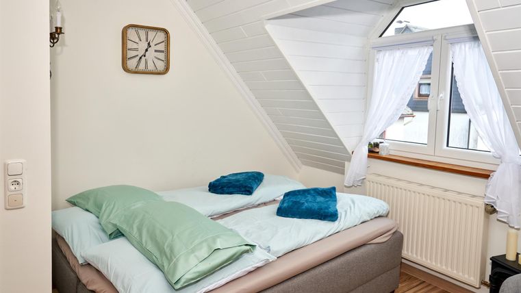 Ein gemütliches Schlafzimmer mit einem Doppelbett und bunten Kissen. Es hat ein schräges Dachfenster und helle Vorhänge, die Licht hereinlassen.