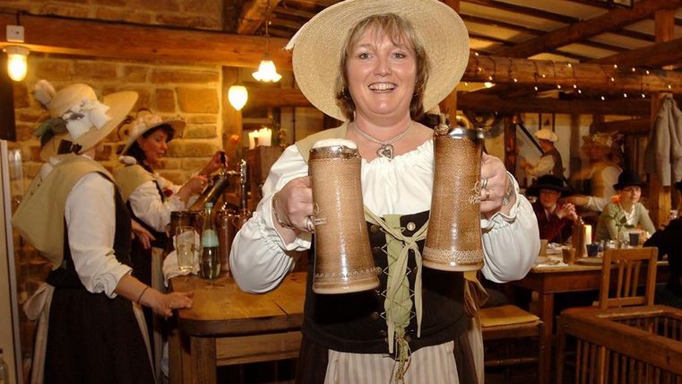 <p>Eine traditionell gekleidete Frau lacht in die Kamera und hält zwei große Bierkrüge in den Händen. Im Hintergrund viele Gäste und weitere Kellnerinnen im Gasthaus der Birgeler Mühle.</p>