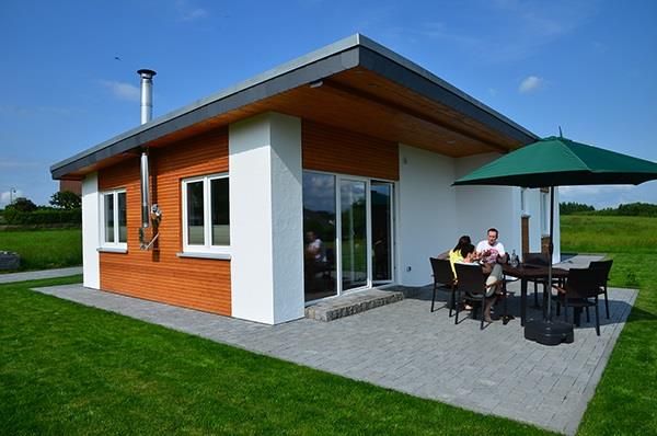Ein modernes Haus mit Holzverkleidung in einer grünen Umgebung. Draußen sitzt eine Gruppe von Menschen bei einem Tisch unter einem Sonnenschirm.