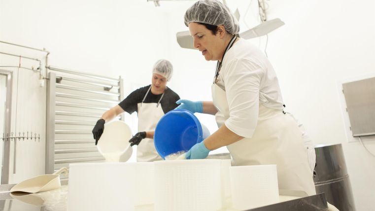 Zwei Personen arbeiten in einer Lebensmittelproduktion. Sie gießen Zutaten in große Behälter und tragen Schutzausrüstung.