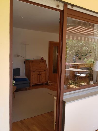 Ein Blick durch die Glastür auf einen hellen Wohnraum. Im Hintergrund sind Möbel und ein Fenster mit Blick nach draußen zu sehen.