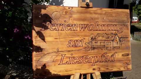 Ein Holzschild mit der Aufschrift "Ferienwohnung am Lieserpfad". Im Hintergrund sind Blumen und eine Wohnsiedlung zu sehen.