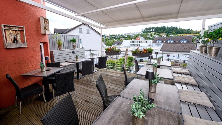 Eine moderne Terrasse mit Holztischen und Stühlen, umgeben von Pflanzen. Im Hintergrund sind Dächer und Hügel zu sehen.