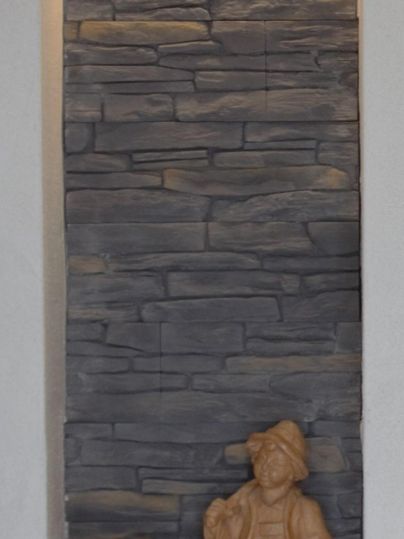 Eine Wand mit einer steinigen Struktur und einem Holzschnitt. Daneben steht eine Holzfigur mit einer wandernden Pose.