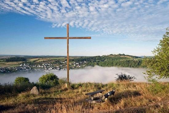 Ein Holzkreuz steht auf einem Hügel mit Blick auf ein Dorf und Nebel im Tal. Der klare Himmel und die sanften Hügel schaffen eine ruhige Atmosphäre.