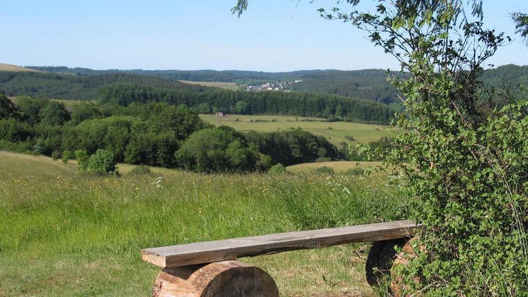 Eine Holzbank steht auf einer Wiese mit Blick auf grüne Hügel und einen kleinen Ort im Hintergrund. Die Umgebung ist ruhig und naturnah.