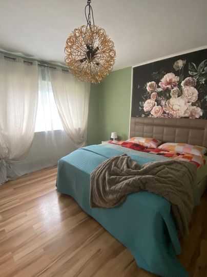 Ein gemütliches Schlafzimmer mit einem großen Bett und einer blauen Decke. Die Wände sind grün mit einer Blumendekoration, und viel natürliches Licht fällt durch die Vorhänge.