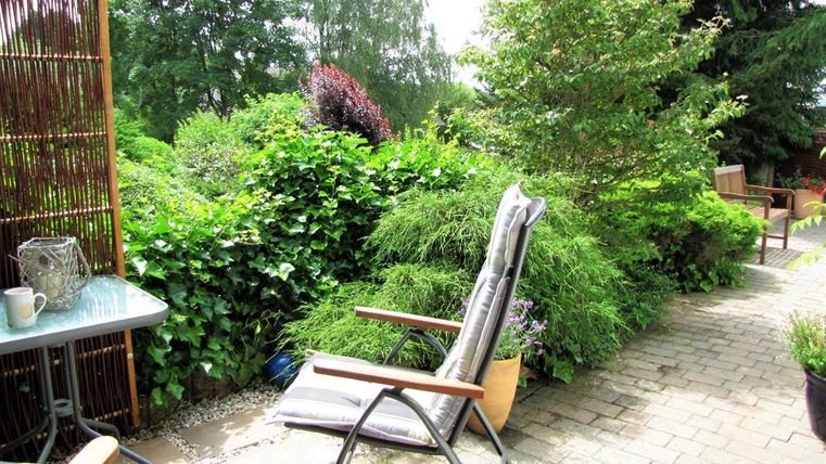 Ein gemütlicher Gartenbereich mit einem Liegestuhl und einem Tisch. Umgeben von üppigem Grün und Pflanzen.