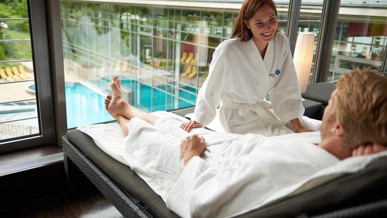 Eine entspannte Atmosphäre in einem Spa-Bereich. Eine Person in einem Bademantel erhält eine Behandlung und lächelt.