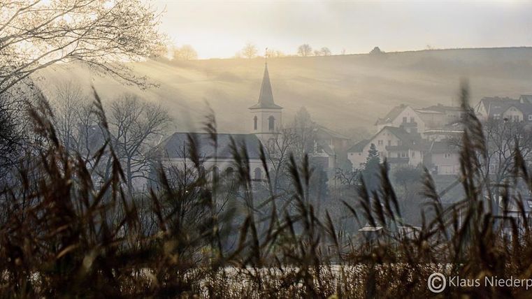 Eine malerische Landschaft mit sanften Hügeln und einem kleinen Dorf im Nebel. Im Vordergrund sind hohe Gräser zu sehen, während ein Kirchturm in der Ferne hervorsticht.